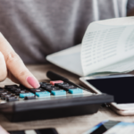 Close-up de uma mão pressionando as teclas de uma calculadora sobre uma mesa. Ao lado, há documentos financeiros, um smartphone e cédulas parcialmente visíveis. A imagem representa controle financeiro, cálculos de despesas, planejamento de orçamento ou gestão de finanças pessoais, com fundo desfocado e iluminação suave em ambiente interno.