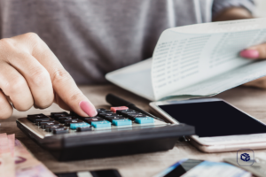 Close-up de uma mão pressionando as teclas de uma calculadora sobre uma mesa. Ao lado, há documentos financeiros, um smartphone e cédulas parcialmente visíveis. A imagem representa controle financeiro, cálculos de despesas, planejamento de orçamento ou gestão de finanças pessoais, com fundo desfocado e iluminação suave em ambiente interno.